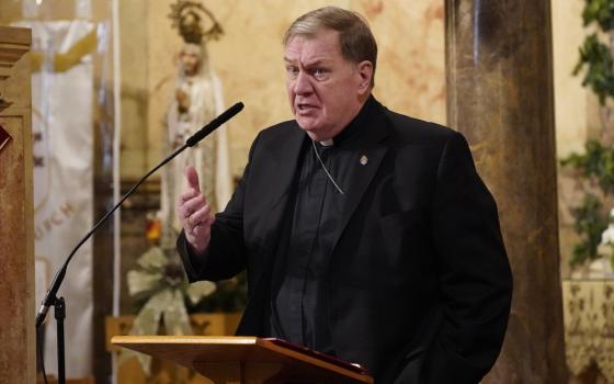 The Cardinal wears plain black clericals, stands at lectern speaking. 