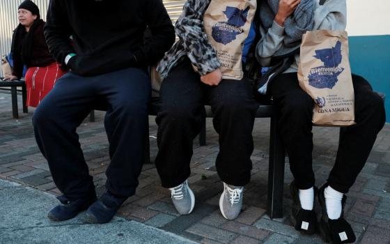 Three people with faces out of frame sit on a bench, two hold paper bags.