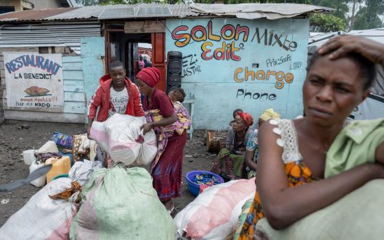 A woman is foregrounded leaning on sacks with hand on head, in background is a small storefront and and other people, including a woman with a baby on her back.