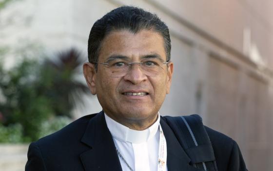 The bishops wears a black suit over white shirt with color, smiles at camera.