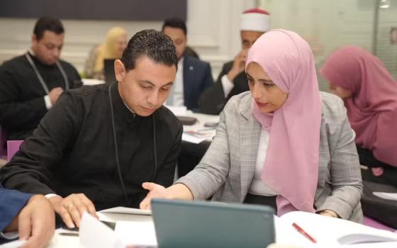 Benadi wearing business garb and pink hijab sits at a table with clergyman explaining concepts.