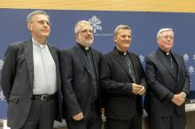 The four men stand in front of a blue and white photo background marked with the synodal insignia.