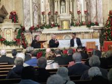 The bishops sits at table in front of altar.